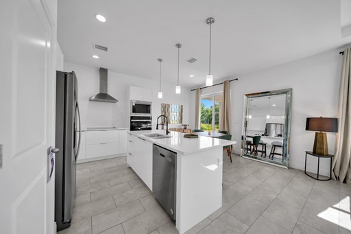 Modern open-plan kitchen and dining area in the Madison model home by Paladium One in LaBelle, Florida, featuring white shaker cabinets, a large island with sink, pendant lighting, stainless steel appliances, and tile flooring.